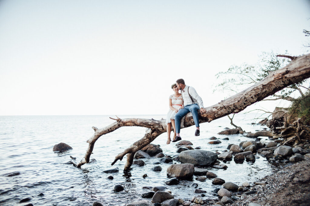 Vintage-Hochzeit auf Fehmarn, Brautpaar am Strand auf einem Baumstamm