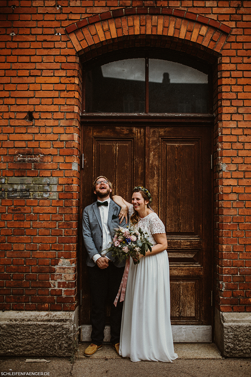 Brautpaar vor Backsteinwand mit üppigem Brautstrauß, sie im Zweiteiler mit Blumenkranz